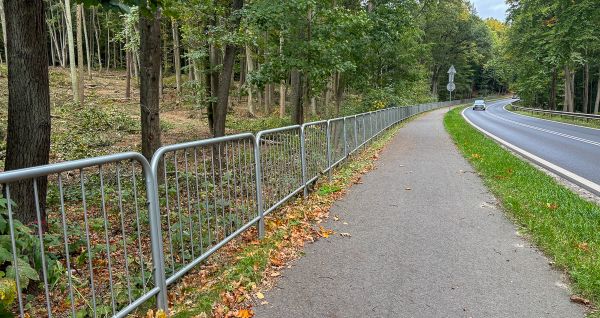 Widok na ścieżkę rowerową biegnącą przez las, po prawej stronie ulica, po lewej barierki i las