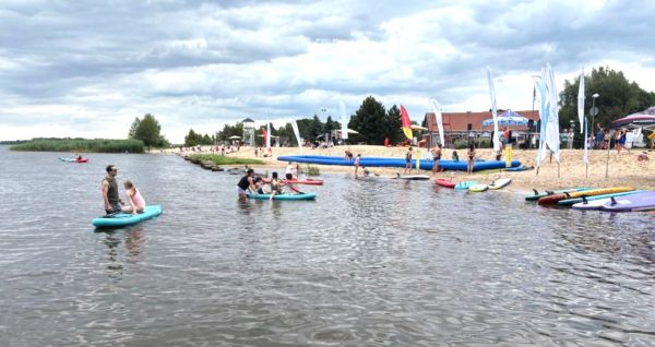 Widok na plażę w Trzebieży od strony wody. Na wodzie osoby pływające na deskach